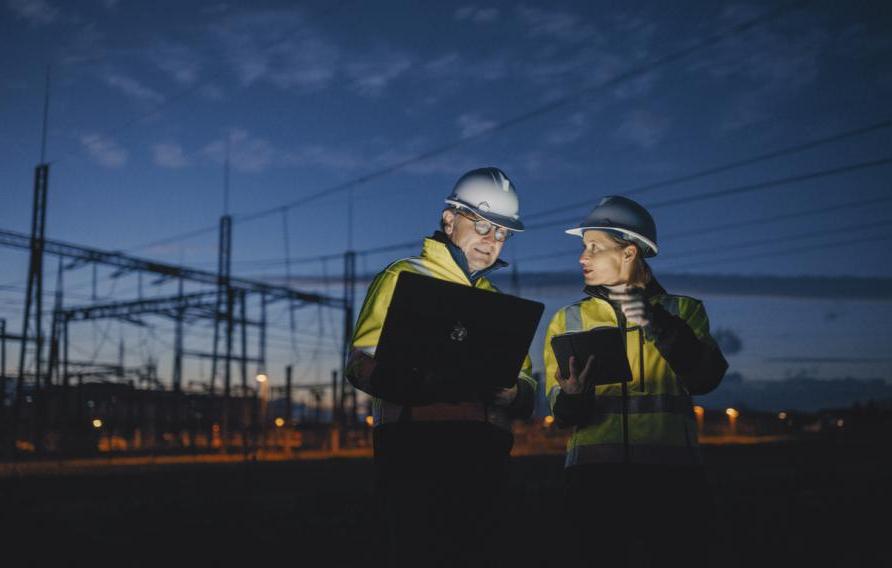 Electrical Engineers Discussing Over Laptop And Digital Tablet While Standing At Dark Power Station During Night stock photo